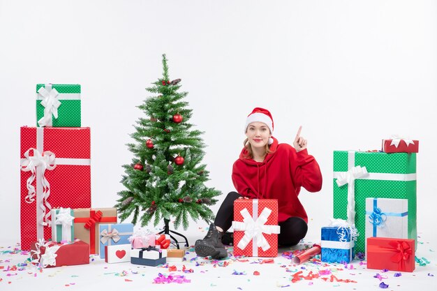 Front view of young woman sitting around holiday presents on white wall