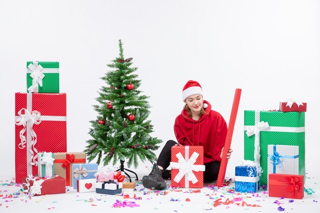 Front view of young woman sitting around holiday presents on white wall