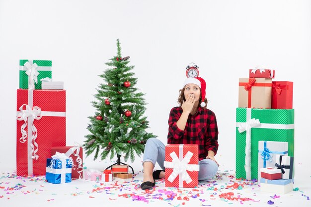 Front view of young woman sitting around presents holding clocks on her head on a white wall