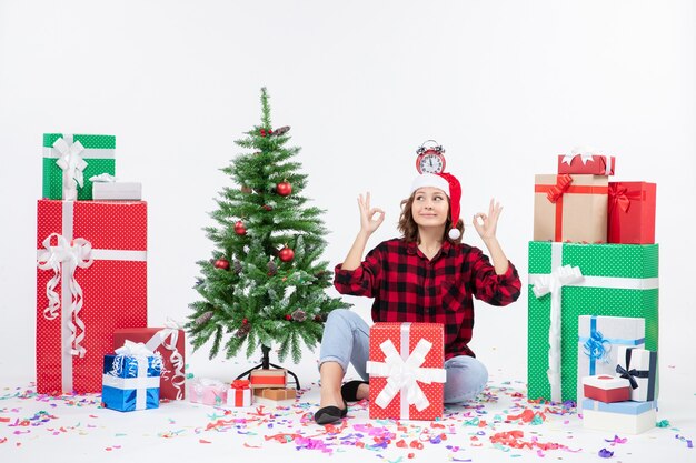 Front view of young woman sitting around presents holding clocks on her head on the white wall