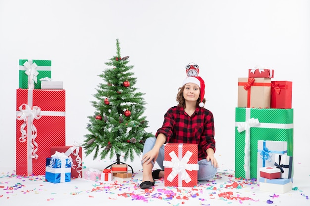 Front view of young woman sitting around presents holding clocks on her head on white wall