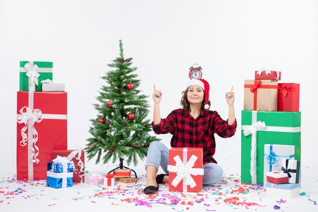 Front view of young woman sitting around presents holding clocks on her head on white wall