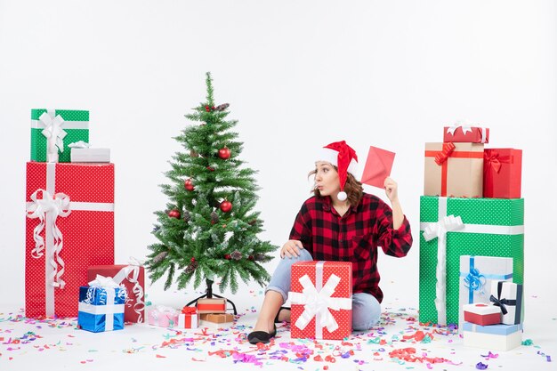Вид спереди молодой женщины, сидящей вокруг подарков, держащей конверт на белой стене
