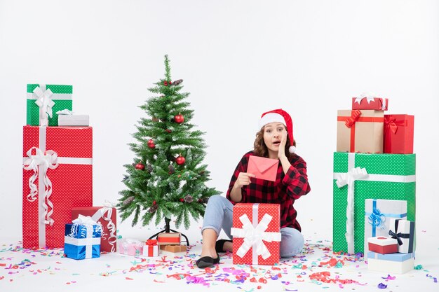 Front view of young woman sitting around presents holding envelop on white wall