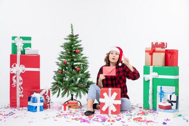 Front view of young woman sitting around presents holding envelop on white wall