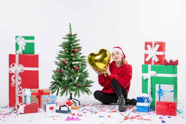 Front view of young woman sitting around presents holding gold heart figure on a white wall