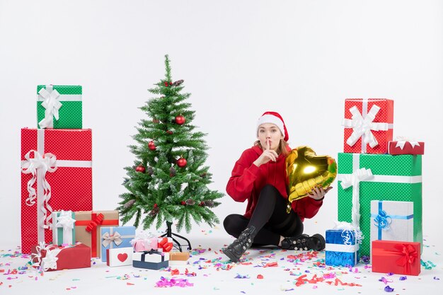 Free photo front view of young woman sitting around presents holding gold heart figure on white wall