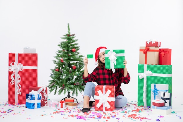 Front view of young woman sitting around presents holding green bank card and present on a white wall