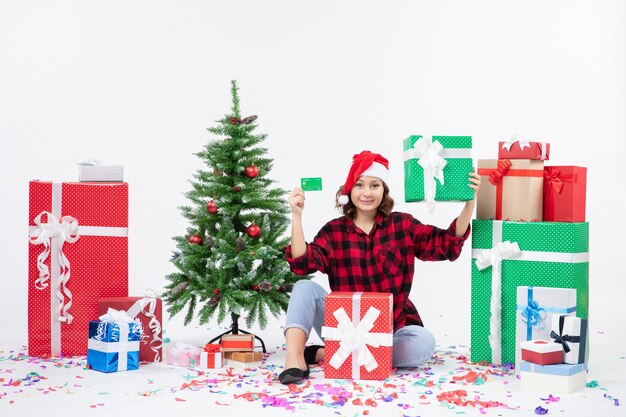 Front view of young woman sitting around presents holding green bank card and present on the white wall