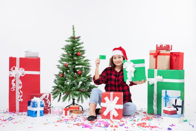 Front view of young woman sitting around presents holding green bank card and present on white wall