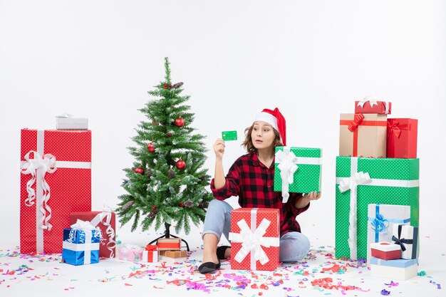 Front view of young woman sitting around presents holding green bank card and present on white wall