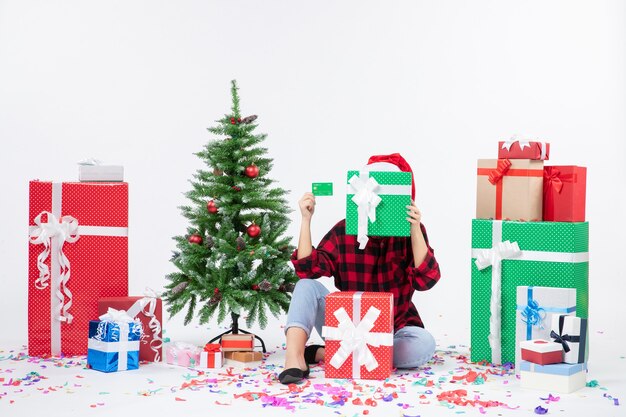 Front view of young woman sitting around presents holding green bank card on a white wall