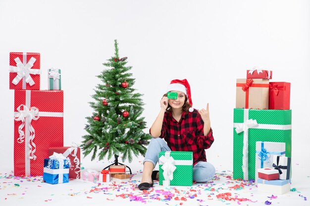 Front view of young woman sitting around presents holding green bank card on a white wall