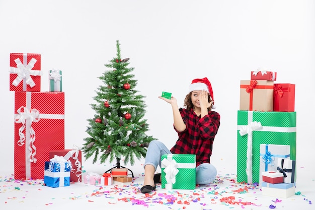 Front view of young woman sitting around presents holding green bank card on a white wall