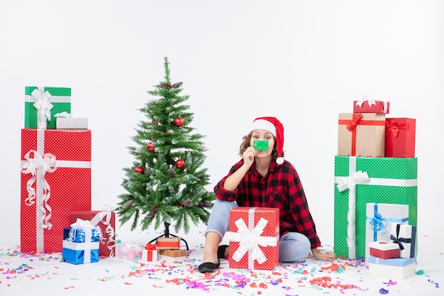 Front view of young woman sitting around presents holding green bank card on a white wall