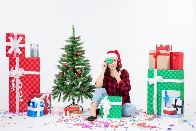 Front view of young woman sitting around presents holding green bank card on the white wall