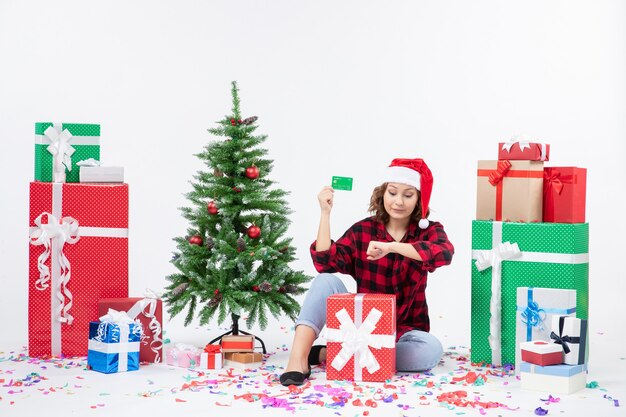 Front view of young woman sitting around presents holding green bank card on white wall