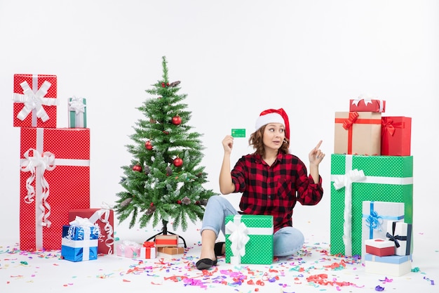 Front view of young woman sitting around presents holding green bank card on white wall