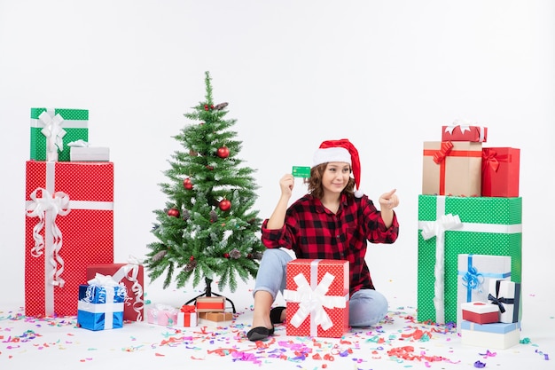 Front view of young woman sitting around presents holding green bank card on white wall