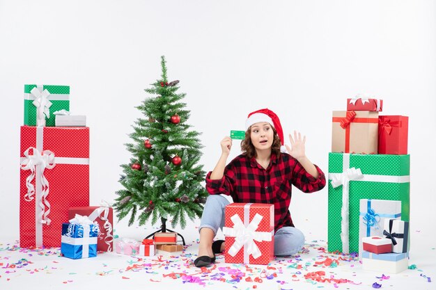 Front view of young woman sitting around presents holding green bank card on white wall