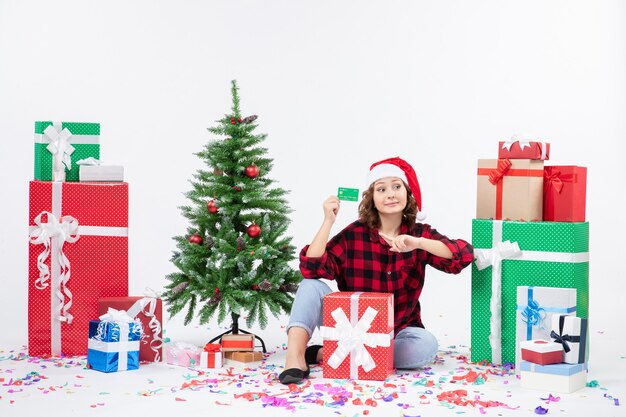 Front view of young woman sitting around presents holding green bank card on white wall