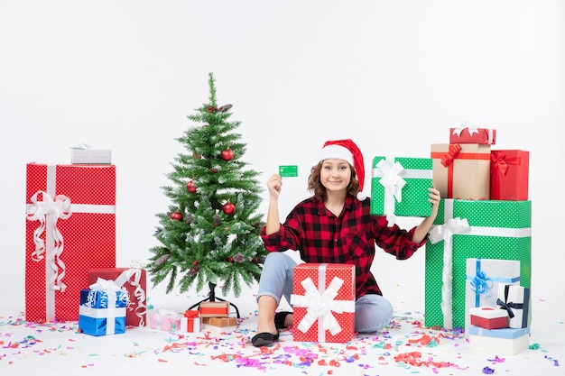 Front view of young woman sitting around presents holding green bank card on white wall