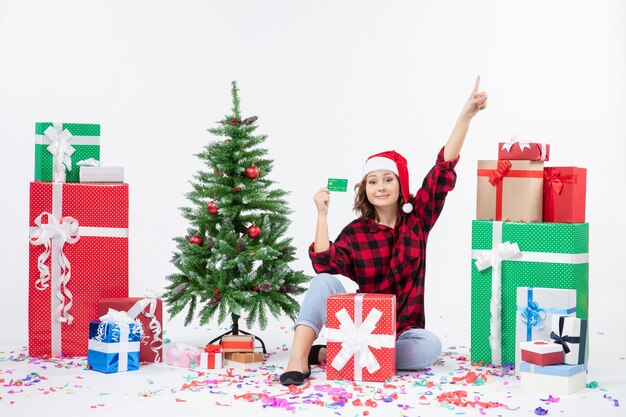 Front view of young woman sitting around presents holding green bank card on white wall