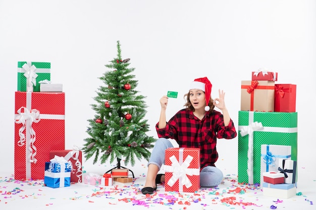 Front view of young woman sitting around presents holding green bank card on white wall