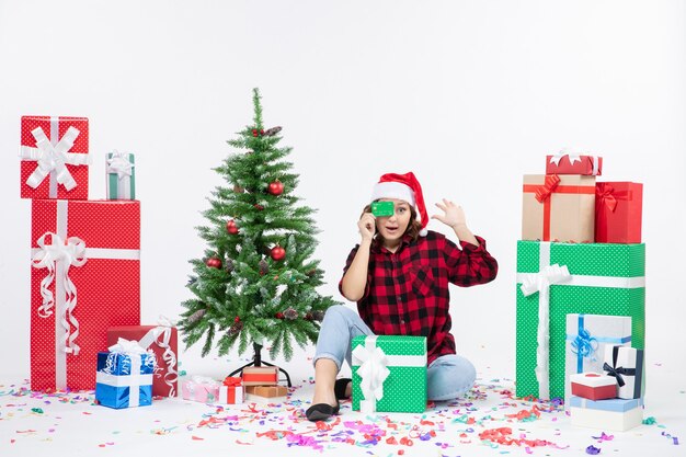 Front view of young woman sitting around presents holding green bank card on white wall