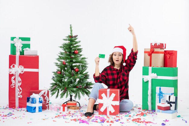 Front view of young woman sitting around presents holding green bank card on white wall