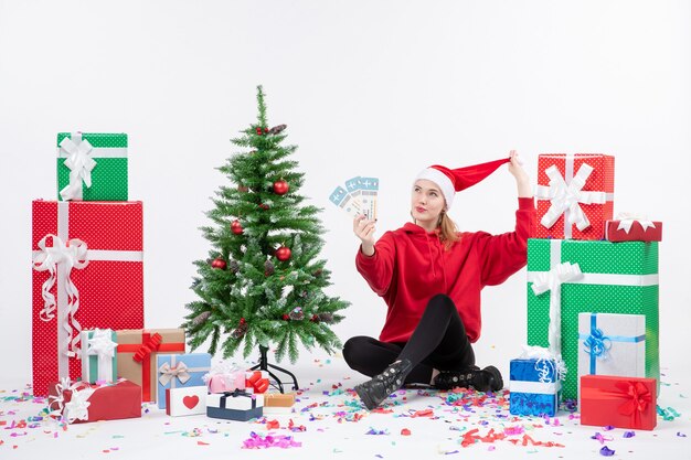 Front view of young woman sitting around presents holding plane tickets on the white wall