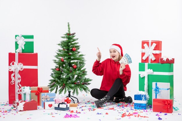 Front view of young woman sitting around presents holding plane tickets on white wall