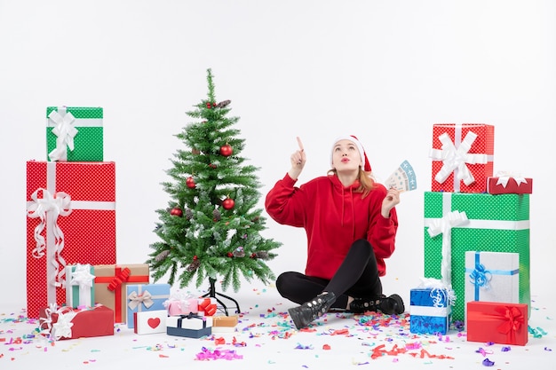 Front view of young woman sitting around presents holding plane tickets on white wall