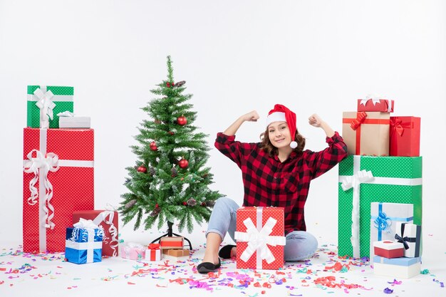 Front view of young woman sitting around presents and little holiday tree on a white wall