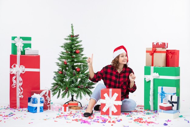 흰 벽에 선물과 작은 크리스마스 트리 주위에 앉아있는 젊은 여자의 전면보기