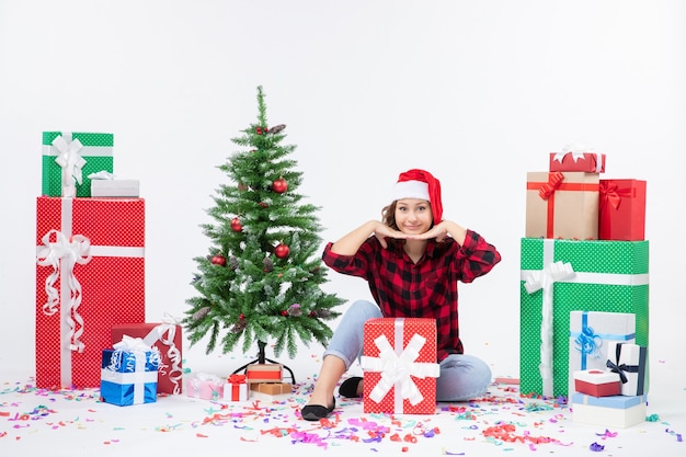 Front view of young woman sitting around presents and little holiday tree on the white wall