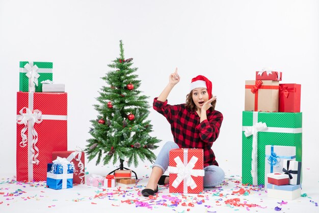 Front view of young woman sitting around presents and little holiday tree on white wall