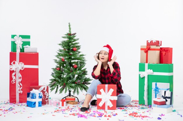 Front view of young woman sitting around presents and little holiday tree on white wall