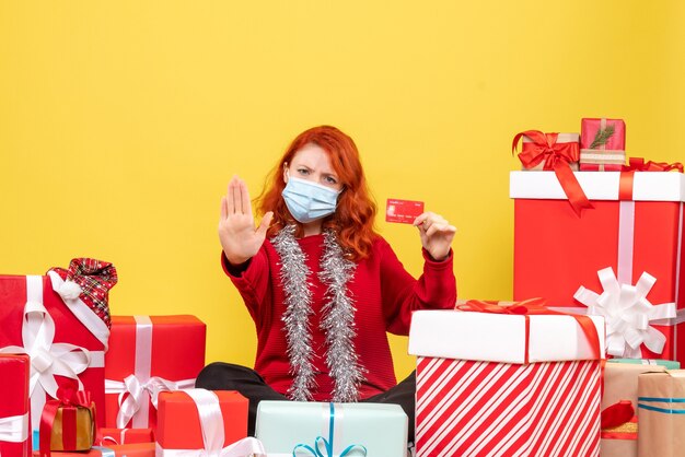 Front view of young woman sitting around presents in mask with bank card on a yellow wall