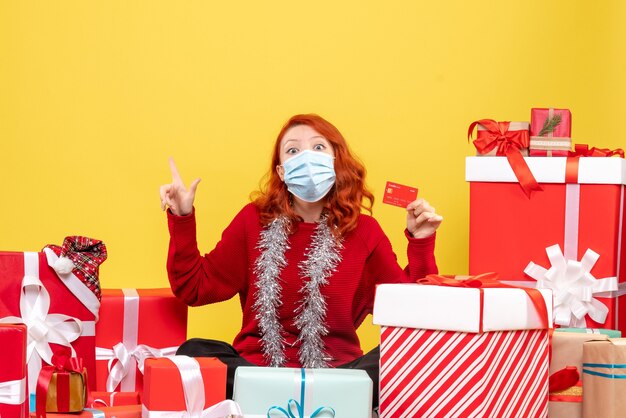 Front view of young woman sitting around presents in mask with bank card on yellow wall