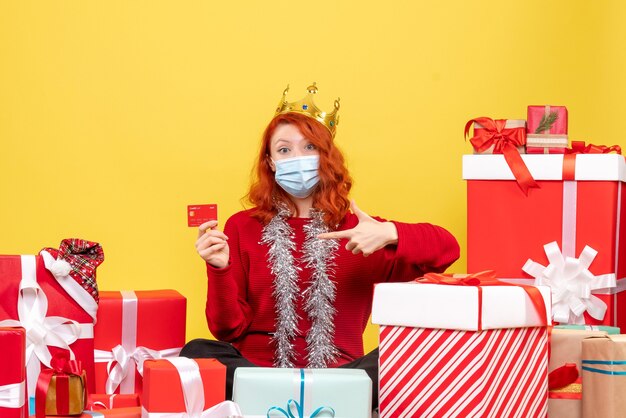 Front view of young woman sitting around presents in mask with bank card on yellow wall