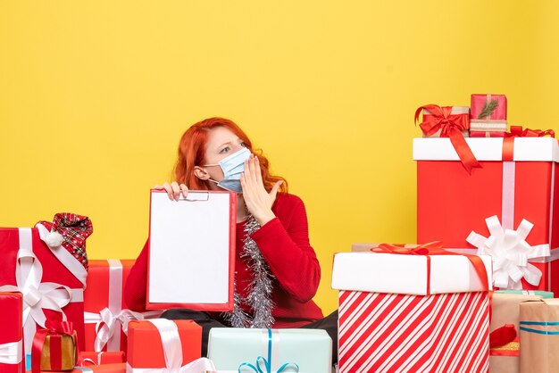 Front view of young woman sitting around presents in mask with file note on yellow wall