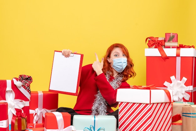 Front view of young woman sitting around presents in mask with file note on yellow wall