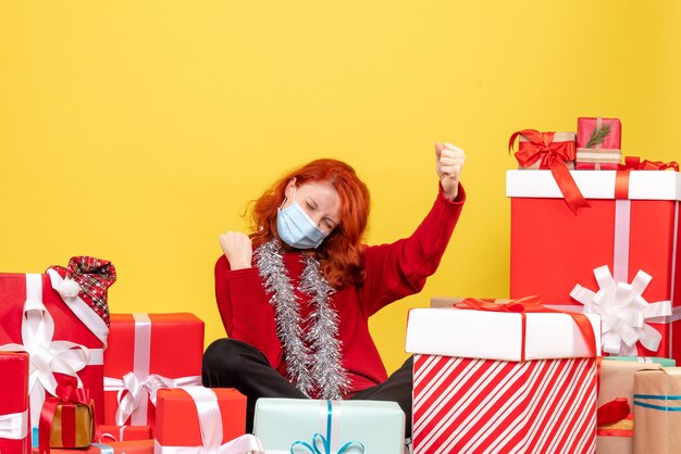 Front view of young woman sitting around presents in mask on a yellow wall