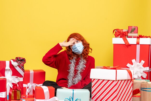 Front view of young woman sitting around presents in mask on the yellow wall