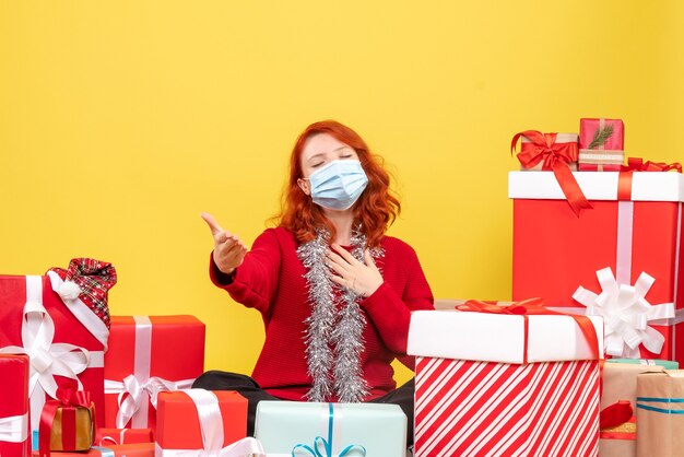 Front view of young woman sitting around presents in mask on the yellow wall