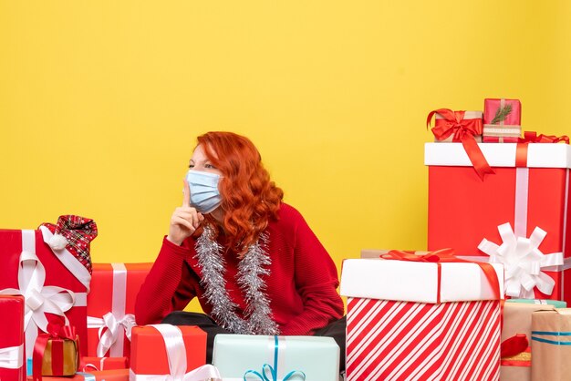 Front view of young woman sitting around presents in mask on the yellow wall