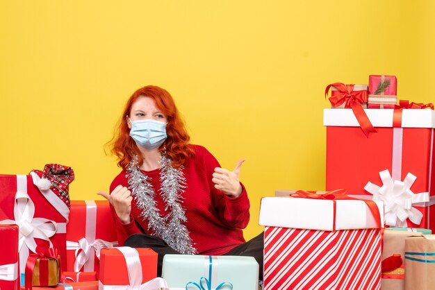 Front view of young woman sitting around presents in mask on yellow wall