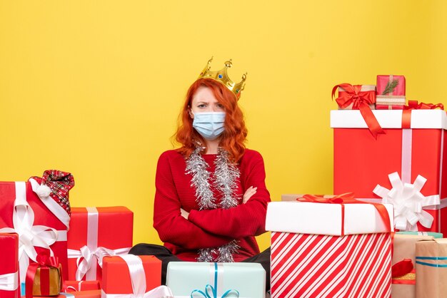 Front view of young woman sitting around presents in sterile mask on yellow wall