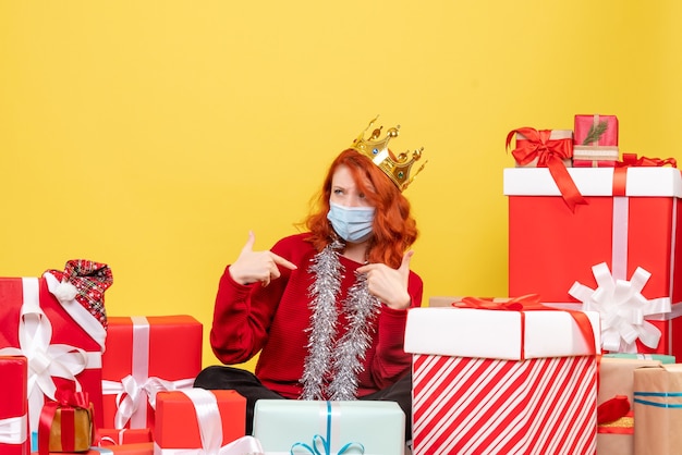 Front view of young woman sitting around presents in sterile mask on yellow wall